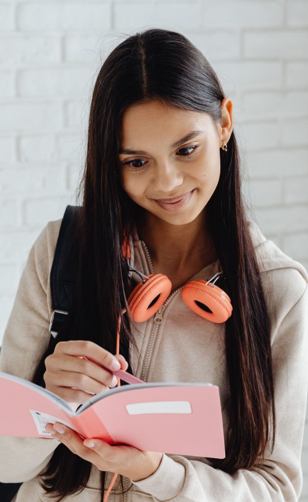 A student in a casual hoodie writing in a notebook, with headphones around her neck